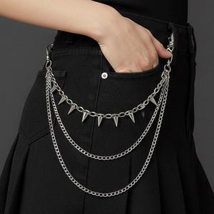 May include: A black pleated skirt with a silver chain belt featuring three strands and cone-shaped spikes. The belt is attached to the skirt's waistband. The skirt is made of a dark fabric and has a classic, punk-inspired design.