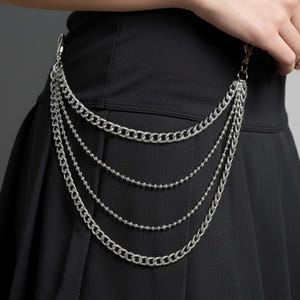 May include: A silver chain belt with multiple strands hangs from a black pleated skirt. The chains are connected to a clasp at the top. The skirt is made of a dark fabric.