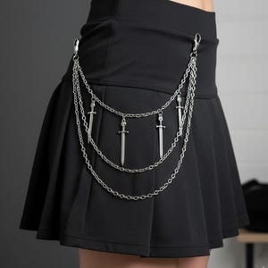 以下が含まれることがあります： 黒のプリーツスカートに、剣のチャームが付いたシルバーチェーンベルト。 スカートはハイウエストでフレアシルエット。 チェーンベルトは3本のストランドで吊り下げられています。