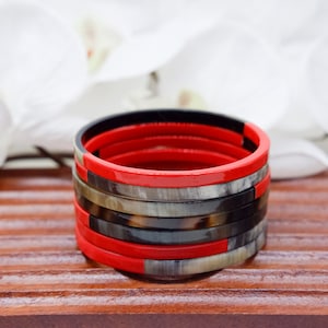 Puede incluir: Pila de pulseras coloridas. Las pulseras son rojas, negras y grises. Las pulseras están apiladas una encima de otra. El fondo está borroso con flores blancas.