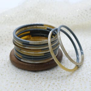 Op de afbeelding: Een stapel kleurrijke armbanden in tinten grijs, geel en wit. De armbanden zijn gestapeld op een kleine, ronde, houten basis. Twee armbanden zijn gescheiden van de stapel, wat de ontwerp- en kleurvariaties laat zien. De achtergrond is een wit oppervlak met gouden glitter.