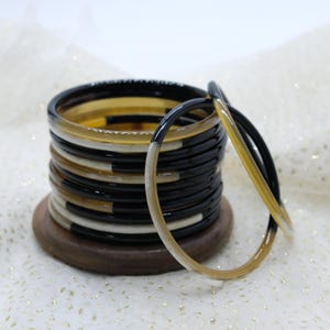 May include: A stack of thin, round bangles in black, cream, and amber hues. The bangles are stacked on a small, round wooden base. Two bangles are separated from the stack, showing the color gradient from black to amber.