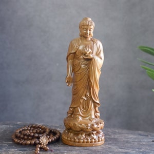May include: A carved wooden Buddha statue in a standing pose, holding a lotus flower. The statue is light brown and stands on a lotus flower base. A strand of wooden prayer beads is in the foreground.