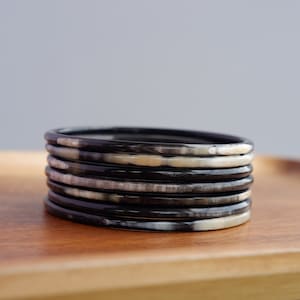 May include: A stack of seven black and cream-colored bangles. The bangles are made of a smooth material and have a slightly rounded rectangular shape. They are stacked on a wooden tray, with a neutral gray background.
