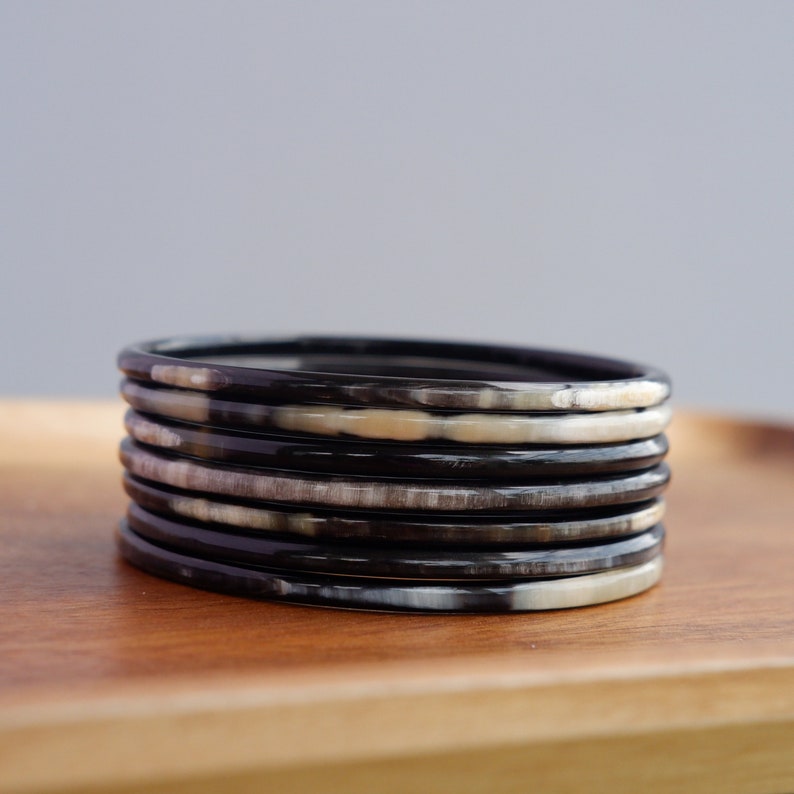 May include: A stack of seven black and cream-colored bangles. The bangles are made of a smooth material and have a slightly rounded rectangular shape. They are stacked on a wooden tray, with a neutral gray background.