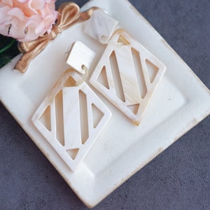May include: A pair of white geometric earrings with a diamond shape and a cut-out design. The earrings are made of a natural material, possibly bone or shell.