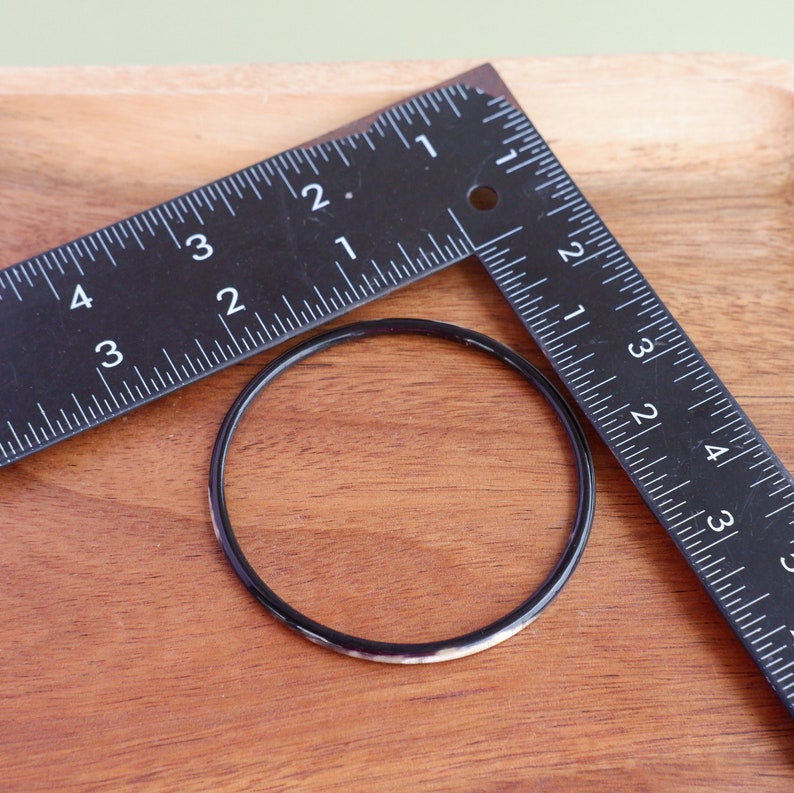 May include: A black bangle bracelet with a white and gray pattern is next to a black ruler on a wooden surface. The ruler is marked with inches. The bangle is approximately 3 inches in diameter.