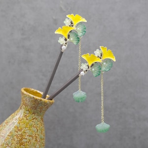 May include: Two gold hair sticks with yellow and green ginkgo leaf designs and dangling green ginkgo leaf charms. The sticks are placed in a brown and yellow ceramic vase.