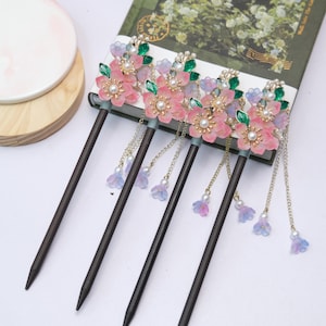 May include: Five wooden hair sticks with pink flower accents, pearl centers, and dangling chains with blue and purple flower charms. The flowers have green leaf accents and are set against a book.