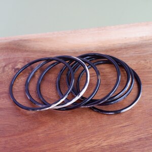 May include: A set of seven black and white bangles. The thin, circular bracelets are made of a dark material with a marbled white and brown pattern. They are arranged on a wooden surface.