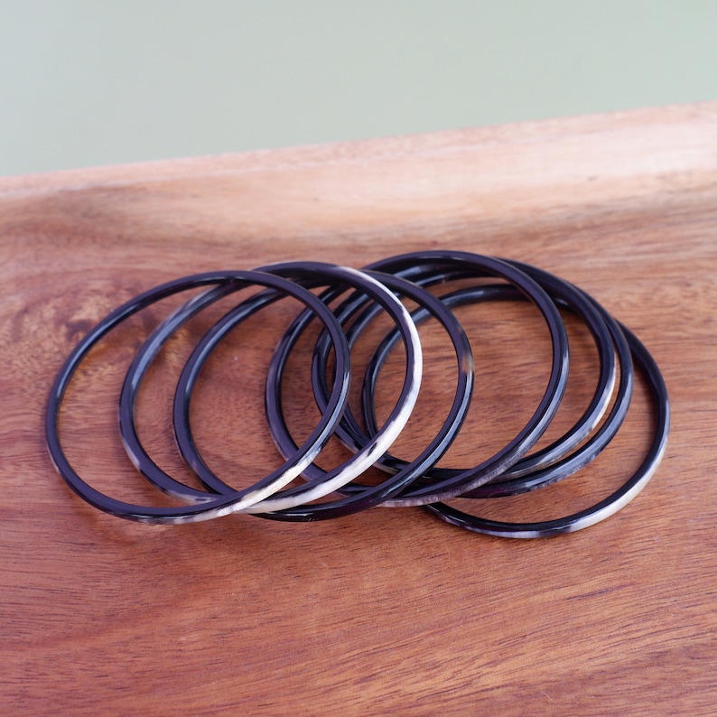 May include: A set of seven black and white bangles. The thin, circular bracelets are made of a dark material with a marbled white and brown pattern. They are arranged on a wooden surface.