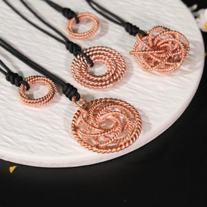 May include: A collection of copper-colored necklaces with black cords. The pendants feature intricate, spiraling designs and circular shapes. The necklaces are displayed on a white, textured surface, showcasing the detailed craftsmanship of each piece.