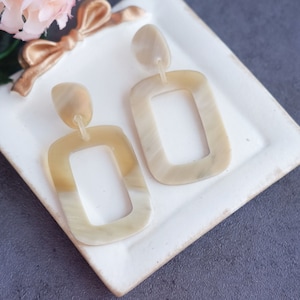 May include: A pair of cream-colored statement earrings with a rectangular shape and a smaller rectangle cut out in the center. The earrings are displayed on a white surface.