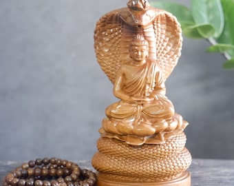 May include: A wooden statue of Buddha seated in meditation, with a cobra hood raised above his head. The statue is carved with intricate details and has a natural wood finish.