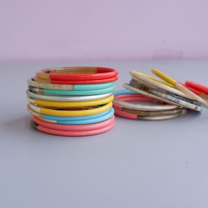 Könnte beinhalten: Ein Stapel bunter Armreifen mit abwechselnden Bändern in Rot, Gelb, Blau und Pink. Die Armreifen sind aus Horn und haben eine natürliche, polierte Oberfläche.