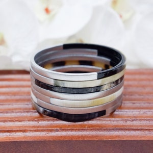 May include: A stack of eight bangles made of horn and metal. The bangles are in black, white, and silver colours. The bangles are stacked on top of each other, creating a layered effect.