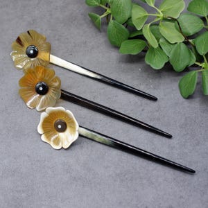 Può includere: Tre bastoncini per capelli marroni e neri con cime a forma di fiore. I fiori sono di colore marrone chiaro e hanno un centro nero.