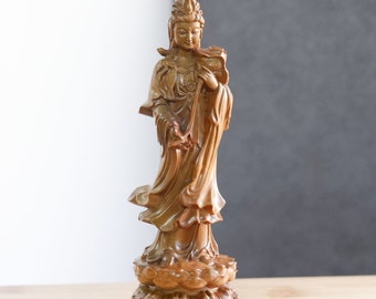 May include: A wooden statue of a female figure with a crown and flowing robes, standing on a lotus base. The statue is carved in a light brown color.