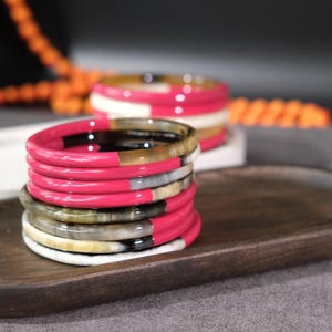 Könnte beinhalten: Ein Stapel bunter Armreifen mit einem leuchtend rosa Akzent. Die Armreifen sind auf einem Holztablett gestapelt, mit einem zweiten Set im Hintergrund. Die Armreifen haben eine Mischung aus Farben, darunter Rosa, Schwarz, Weiß und Grau.