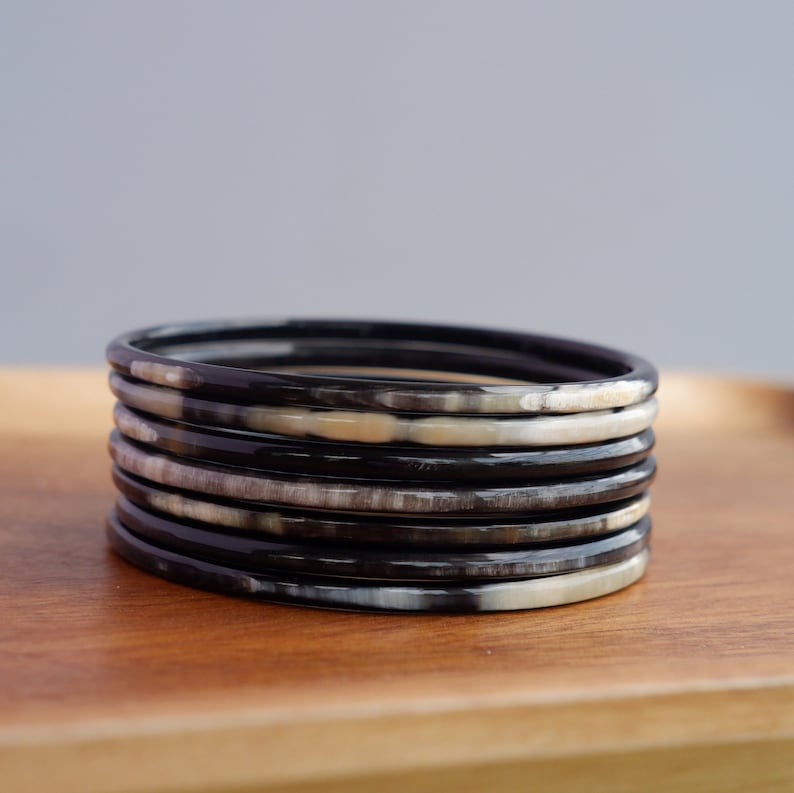 May include: Stack of seven black and cream-colored bangles. The bracelets are made of a glossy material and are stacked on top of each other. The bangles are on a wooden surface.