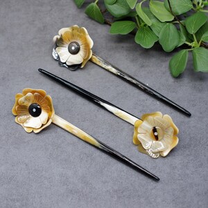 Può includere: Tre bastoncini per capelli con cime a forma di fiore. I bastoncini sono fatti di corno marrone e nero e i fiori sono fatti di un materiale marrone chiaro con un centro marrone scuro.