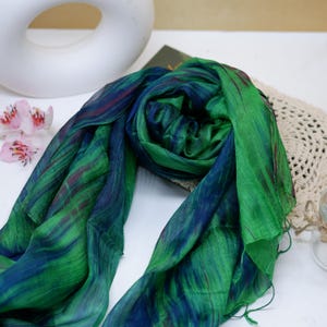 May include: A lightweight scarf featuring a vibrant blend of emerald green and deep navy blue colors with subtle streaks of dark purple. The fabric has a soft, semi-sheer texture with delicate frayed edges, creating a fluid and draped appearance.