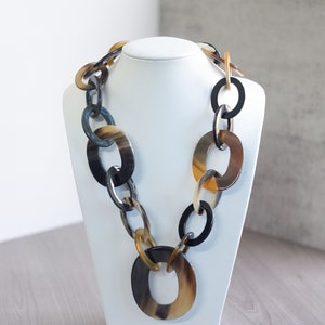 Puede incluir: Un collar largo hecho de grandes eslabones ovalados de cuerno en tonos marrones, negros y beige. El collar se muestra en un busto de maniquí blanco.