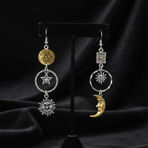 May include: A pair of celestial-themed earrings. One earring features a gold-toned moon disc, a silver star within a ring, and a sun pendant. The other earring has a star-shaped charm, a sunburst within a ring, and a gold crescent moon.