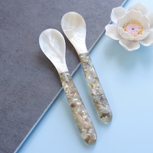 May include: Two white mother of pearl spoons with iridescent handles. The spoons are on a gray and blue surface.