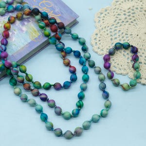 Puede incluir: Un collar largo y una pulsera a juego hechos de cuentas redondas cubiertas de tela. Las cuentas presentan un vibrante patrón tie-dye multicolor en tonos azul verdoso, azul, morado y naranja, con cada cuenta separada por un pequeño nudo.