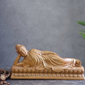 May include: A wooden sculpture of a reclining Buddha, carved in detail, lying on a platform with intricate carvings. The sculpture is a light brown color.