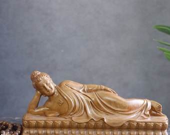 May include: A wooden sculpture of a reclining Buddha, carved in detail, lying on a platform with intricate carvings. The sculpture is a light brown color.