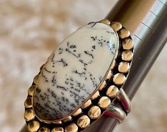 May include: A silver ring with a white and black dendritic agate stone set in a gold beaded bezel. The ring is sized on a metal ring sizer.