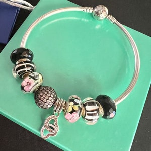May include: A silver bangle bracelet with various decorative beads. The beads include black faceted beads, striped beads, floral beads with pink flowers, and a bead with a diamond-like pattern. A silver charm with two connected hearts is also attached.