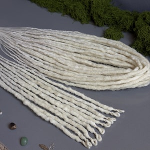 May include: Off-white dreadlocks arranged on a gray surface, with a bundle of the same color in the background. The dreadlocks are thick and textured, with a natural, handmade appearance. Green moss is visible in the background.