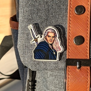 May include: A clear acrylic pin featuring a detailed illustration of Geralt of Rivia, a fictional character from the Witcher series. Geralt is depicted with his silver sword, a blue and black tunic, and a silver medallion. The pin is attached to a gray backpack.