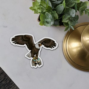 May include: A sticker featuring a brown and white bald eagle with its wings spread, holding a gold and blue helmet in its talons.