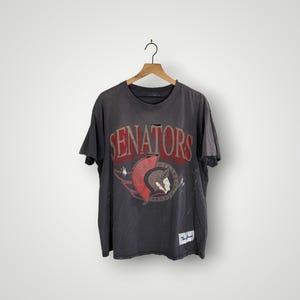 May include: A charcoal gray t-shirt with the word "SENATORS" in red block letters. The shirt features a distressed graphic of a gladiator and a red emblem. The shirt is hanging on a wooden hanger.