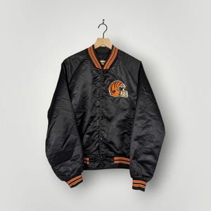 May include: A black satin bomber jacket featuring a Cincinnati Bengals football helmet logo on the left chest. The jacket has a snap-button front closure, long sleeves with ribbed cuffs, and a ribbed collar with orange and black striped detailing.