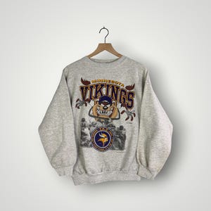 Puede incluir: Sudadera gris claro de los Minnesota Vikings con un gráfico del logotipo y la mascota del equipo. La sudadera tiene mangas largas y cuello redondo. El nombre del equipo se muestra en morado y dorado.