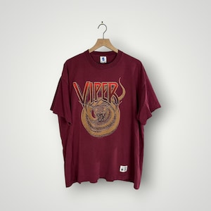 Könnte beinhalten: Ein weinrotes T-Shirt mit einer Grafik einer zusammengerollten Schlange und dem Wort "VIPER" in roten und goldenen Buchstaben. Das T-Shirt hängt an einem Holzbügel vor weißem Hintergrund. Unten am Shirt ist ein kleines Etikett zu sehen.