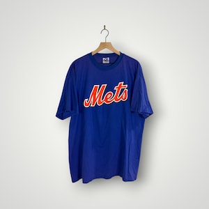 Puede incluir: Una camiseta azul de los Mets con la palabra "Mets" en escritura naranja con un contorno blanco. La camiseta está colgada en una percha de madera sobre un fondo blanco.
