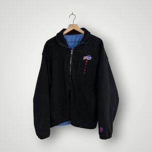 Könnte beinhalten: Schwarze Jacke mit Reißverschluss und blauem Innenfutter. Die Jacke hat das Buffalo Bills-Logo auf der Brust und das Wort "BILLS" in Rot. Ein lila "P" ist auf dem Ärmel gestickt. Die Jacke hängt an einem Holzbügel.
