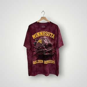 May include: Maroon tie-dye t-shirt with the words "MINNESOTA GOLDEN GOPHERS" in gold lettering. The shirt features a graphic of a football helmet. The shirt is hanging on a wooden hanger.