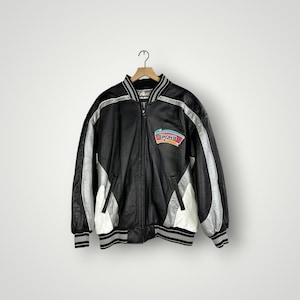 May include: Black and silver varsity jacket with a full zip closure. The jacket features a team logo patch on the chest and white and black striped trim on the collar, cuffs, and hem. The jacket is hanging on a wooden hanger.