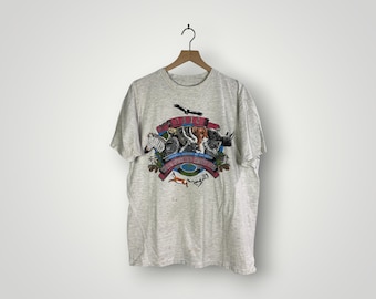 May include: A light gray t-shirt featuring a colorful graphic of various African animals, including a zebra, rhino, and elephant. The graphic is framed by a banner with the word "Kenya" and the phrase "The Heart of Africa".