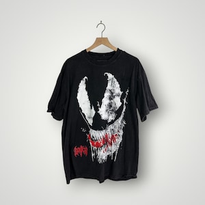 May include: Black t-shirt with a white and red Venom graphic. The design shows a white face with sharp teeth and a red tongue. The word "Venom" is printed in red at the bottom of the design.