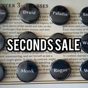 Può includere: Una collezione di bottoni rotondi neri e argentati con diverse classi di personaggi di Dungeons and Dragons stampate su di essi, tra cui Druido, Paladino, Ladro, Monaco e Stregone. I bottoni sono disposti su una superficie bianca con testo sulle classi sullo sfondo. Il testo recita "SECONDS SALE".