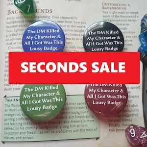 May include: Four green, blue, red and maroon button badges with the text "The DM Killed My Character & All I Got Was This Lousy Badge". The badges are arranged on a table with a white background. The text "SECONDS SALE" is written in red letters on a white background.