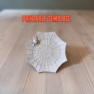 Op de afbeelding: Een witte, achthoekige plaat met een spinnenwebontwerp en een kleine spin. De oranje tekst erboven luidt "PRINTABLE TEMPLATE". De plaat staat op een houten oppervlak.